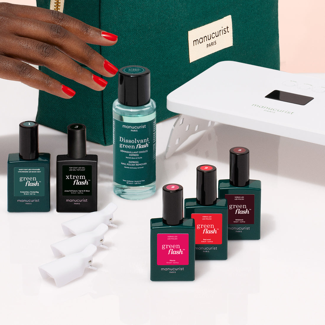 A hand with red nails reaches for the Manucurist Starter Kit, featuring all essentials for an ultra-glossy manicure—vitaminized base coat, polish remover, clips, UV lamp, and a green Paris pouch—arranged on a white surface.