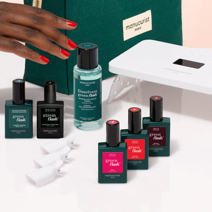 A hand with red nails reaches for the Manucurist Starter Kit, featuring all essentials for an ultra-glossy manicure—vitaminized base coat, polish remover, clips, UV lamp, and a green Paris pouch—arranged on a white surface.