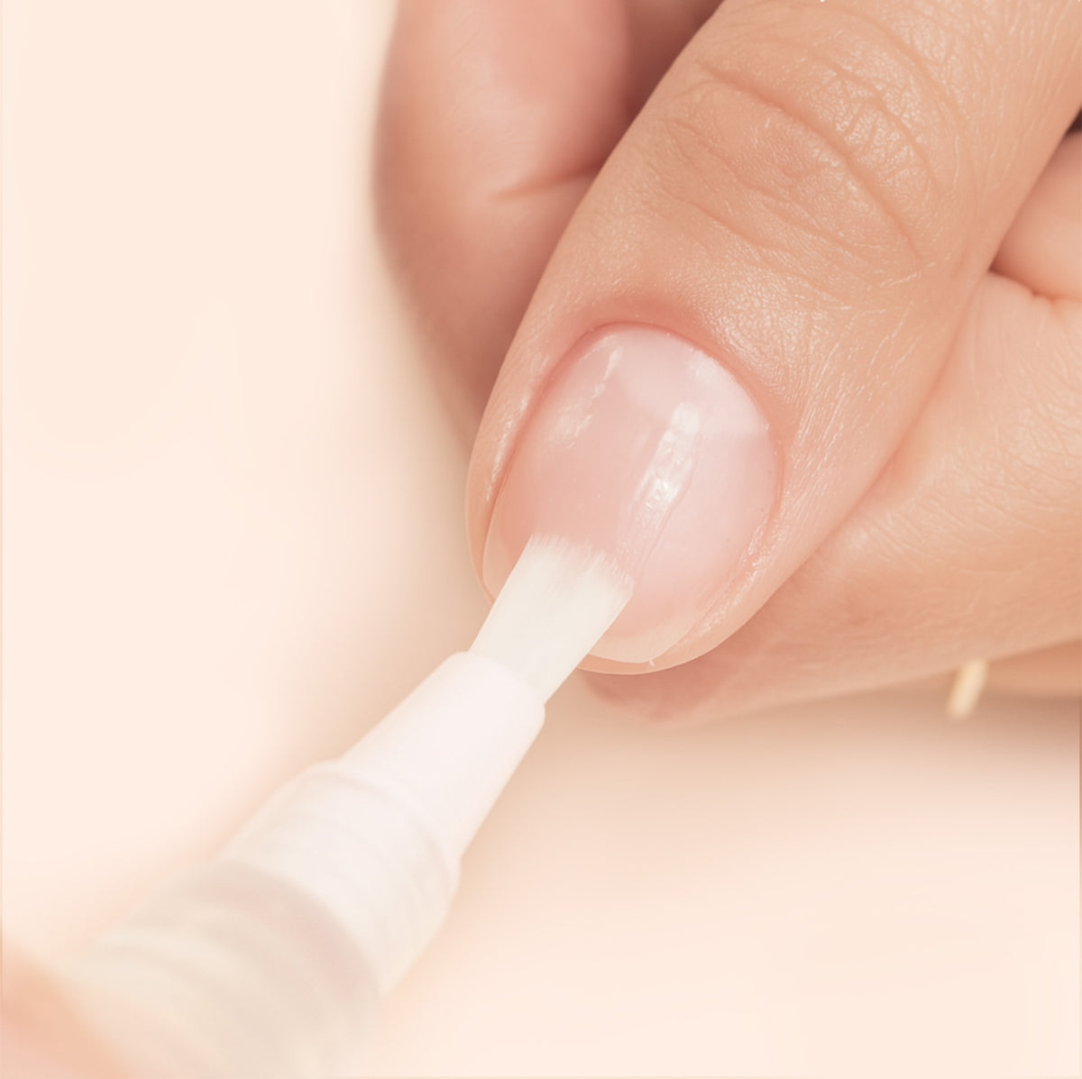 A close-up of a hand applying clear nail polish to a fingernail with a white brush applicator. The background is a soft beige color.