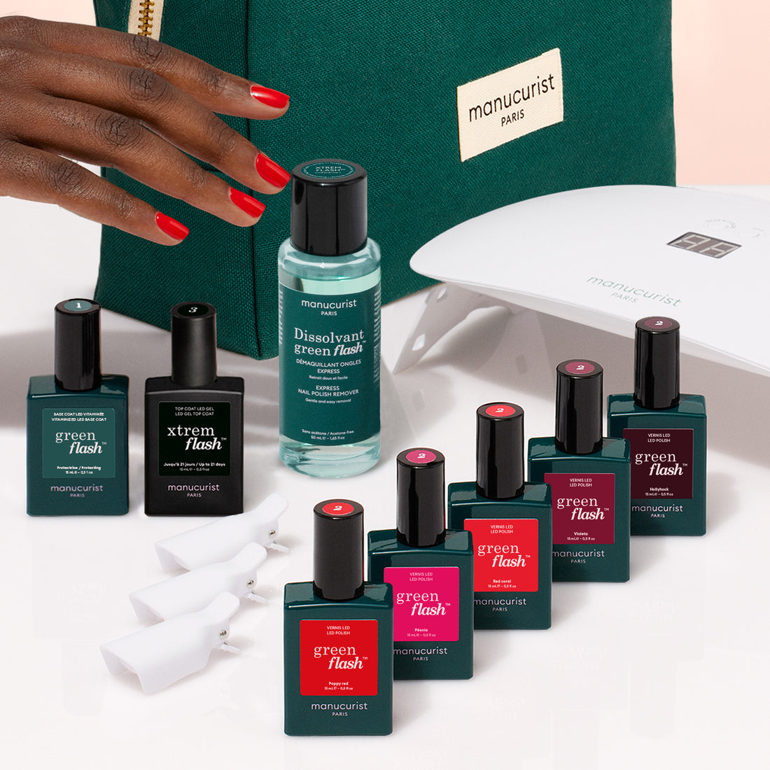 A hand with red nails reaches for a green Manucurist bag. In front are items from the Manucurist Super Starter Kit: Green Flash gel polish, remover, a black bottle, nail clips, and a white LED lamp for a lasting manicure.