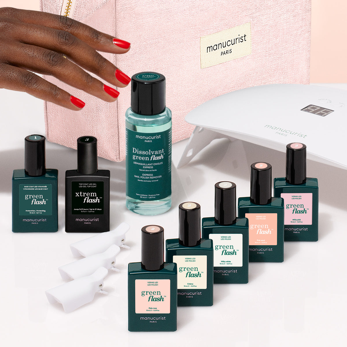 A hand with bright red nails hovers over a Manucurist Super Starter Kit - Soap Nails LED Gel Polish, which includes an LED lamp, remover, and seven colorful bottles arranged in front of a pink cosmetic bag for an ultra-glossy manicure.