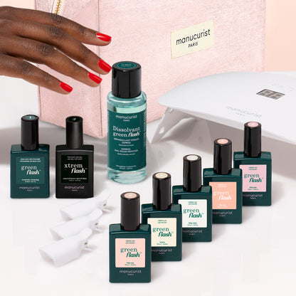A hand with bright red nails hovers over a Manucurist Super Starter Kit - Soap Nails LED Gel Polish, which includes an LED lamp, remover, and seven colorful bottles arranged in front of a pink cosmetic bag for an ultra-glossy manicure.