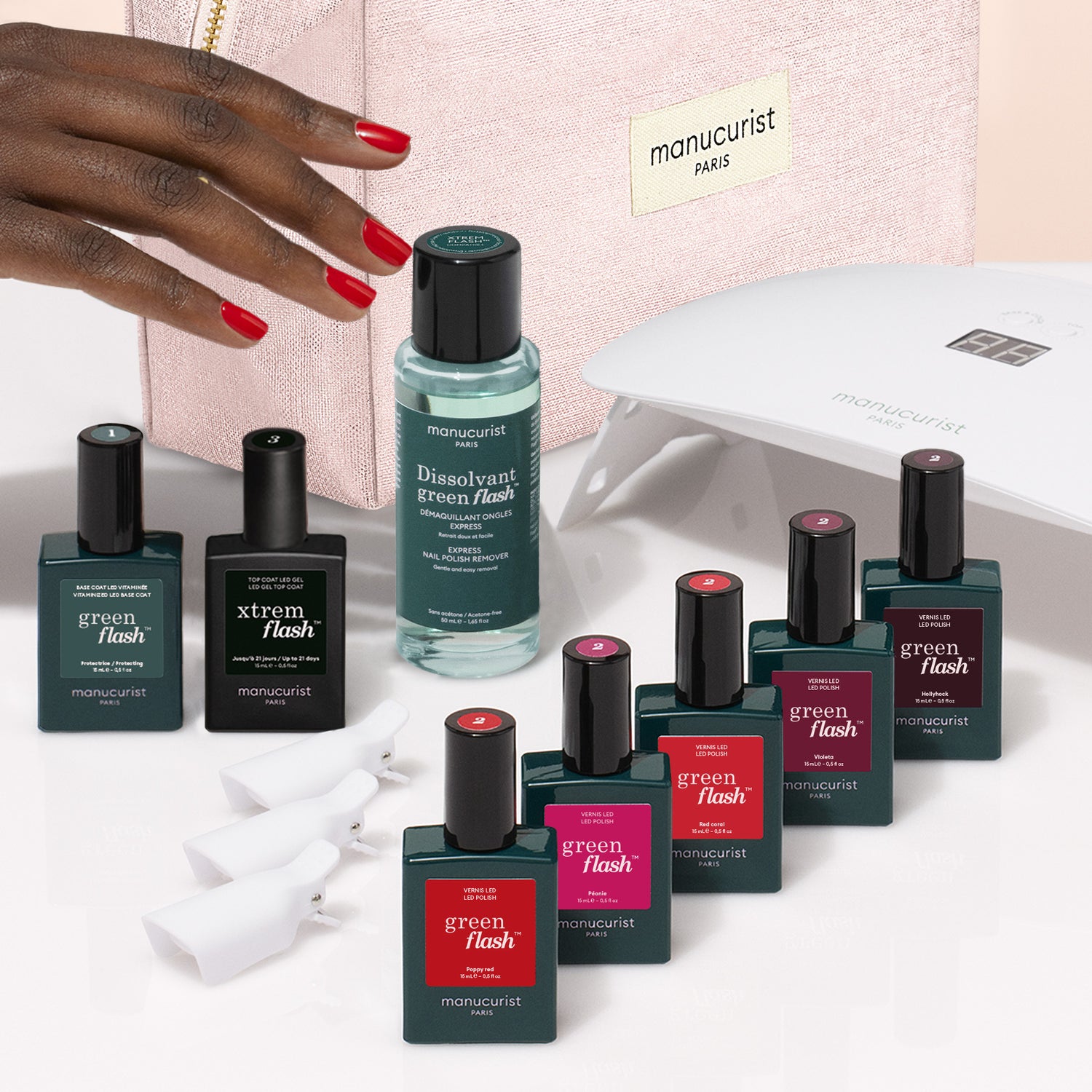 A hand with red nails rests near bottles from the Manucurist Super Starter Kit, including gel polishes, nail polish remover, a UV lamp, removal clips, and a pink bag. The products are arranged on a white surface, promising an ultra-glossy manicure that lasts.