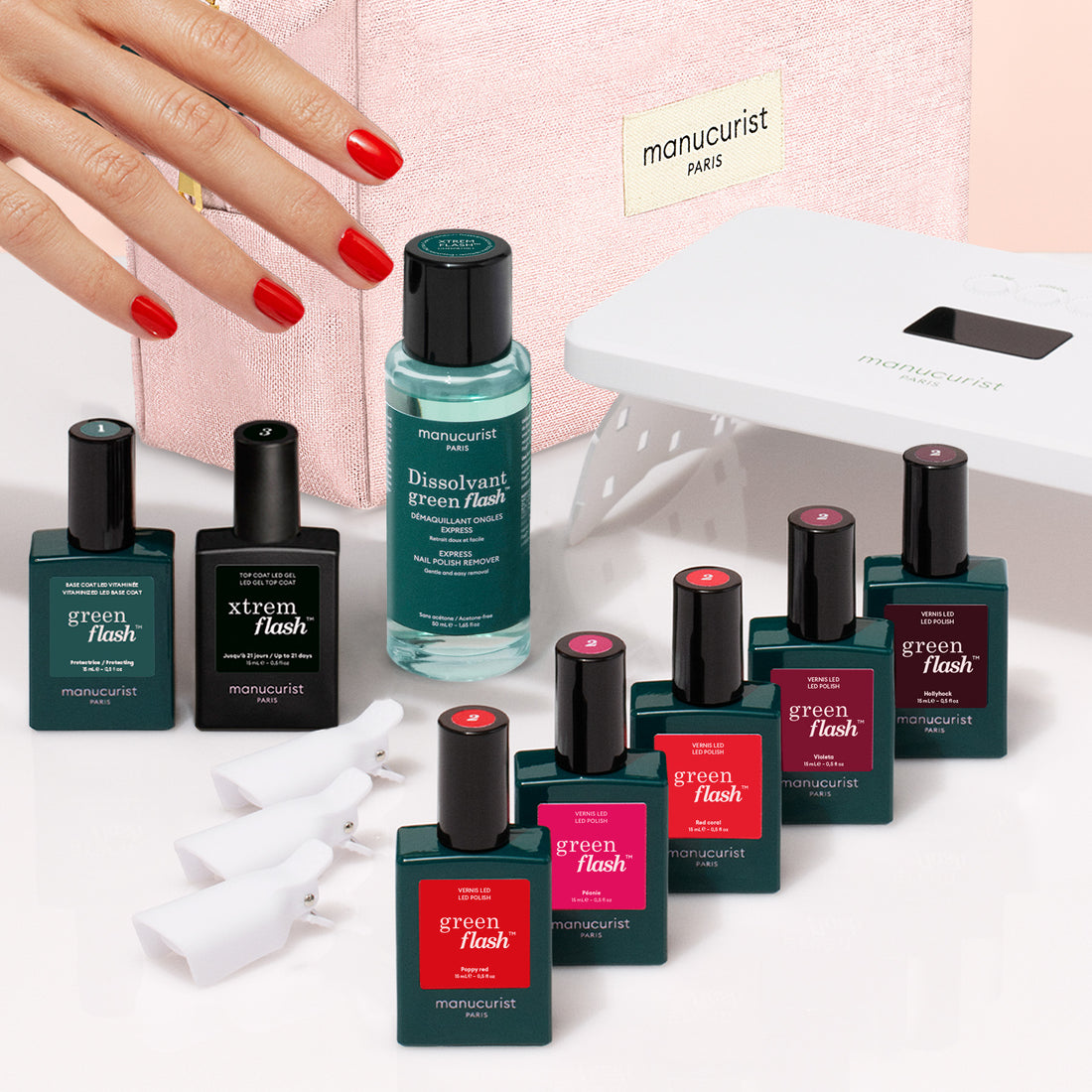 A hand with red nails rests near bottles of Manucurist products from the Super Starter Kit, including nail polish, remover, a UV lamp, and white nail clips—all arranged on a pink surface with a pink Manucurist case in the background for an ultra-glossy manicure.