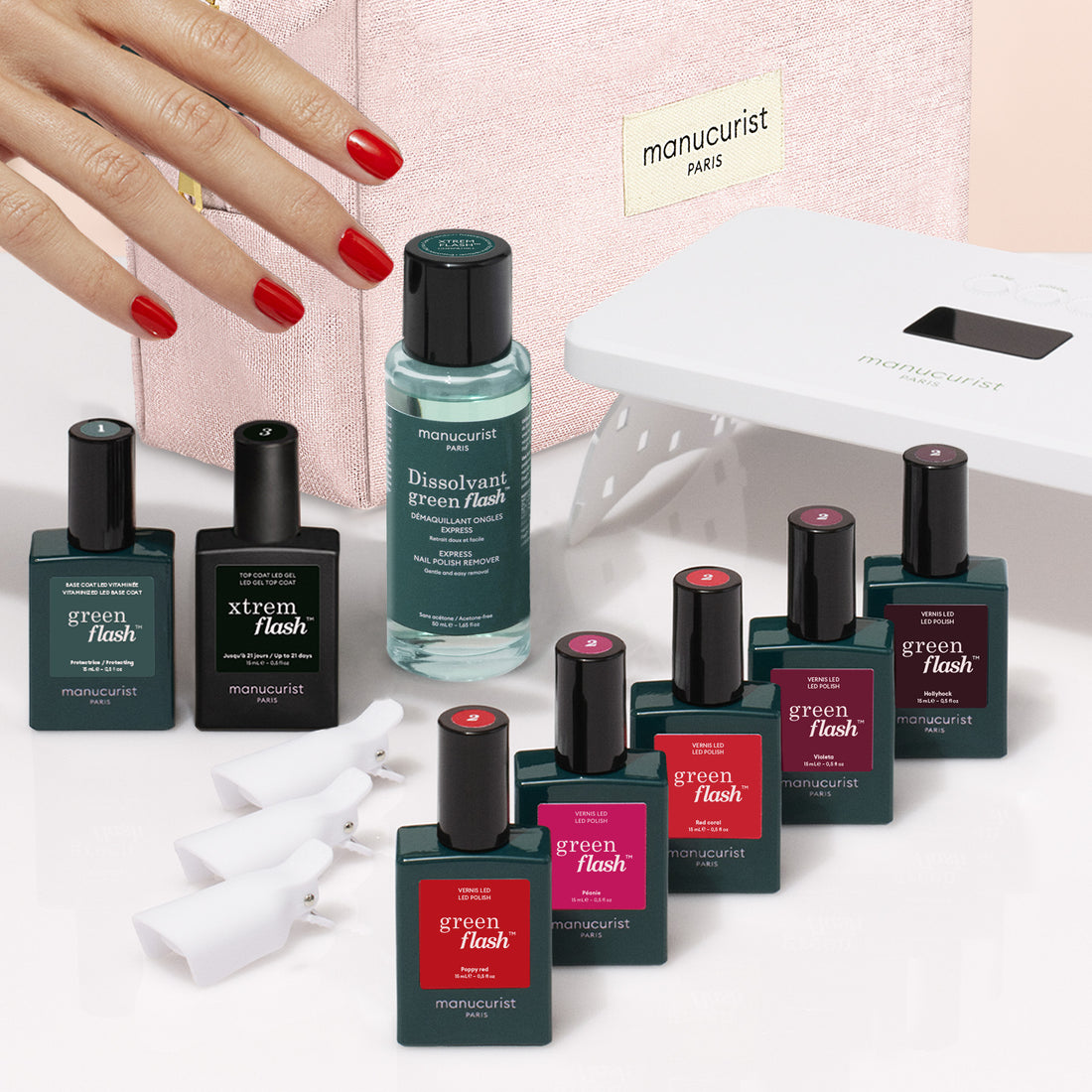 A hand with red nails rests near bottles of Manucurist products from the Super Starter Kit, including nail polish, remover, a UV lamp, and white nail clips—all arranged on a pink surface with a pink Manucurist case in the background for an ultra-glossy manicure.