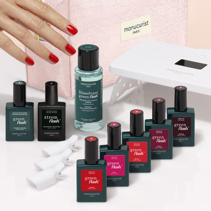 A hand with red nails rests near bottles of Manucurist products from the Super Starter Kit, including nail polish, remover, a UV lamp, and white nail clips—all arranged on a pink surface with a pink Manucurist case in the background for an ultra-glossy manicure.