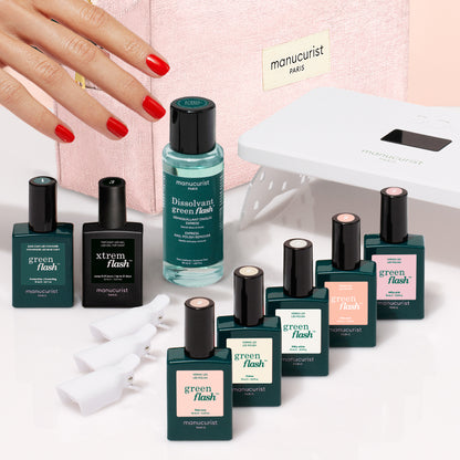 A hand with red-painted nails reaches toward a Super Starter Kit - Soap Nails LED Gel Polish, with bottles, remover, an LED lamp, and clips arranged in front of a pink cosmetic bag to achieve an ultra-glossy manicure.