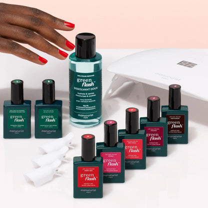 A hand with red polished nails reaches toward the Manucurist Super Starter Kit—featuring long-lasting Green Flash gel polish, remover, a UV lamp, and nail clips—everything needed for an ultra-glossy manicure on a white surface.