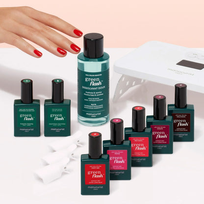 A hand with red polished nails reaches toward the Manucurist Super Starter Kit, featuring Green Flash gel polish, remover, cotton clips, and a UV nail lamp—all arranged on a light surface for an ultra-glossy manicure.