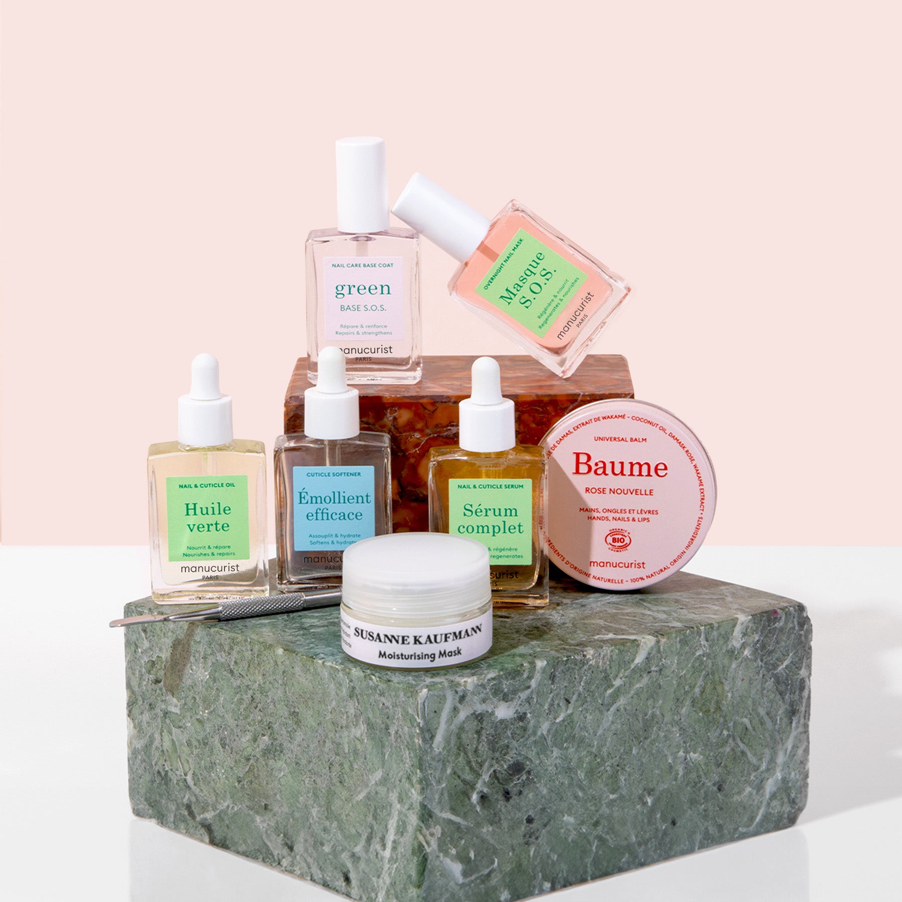 A collection of skincare products, including bottles, a jar, and a tin, arranged on stacked stone blocks with a neutral pink background. Labels on products display names like Manucurist and Susanne Kaufmann.