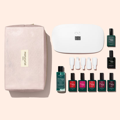 The Manucurist Super Starter Kit features a pink zippered pouch, a white LED nail lamp, green and red gel polish bottles, a remover bottle, and four white nail tools—all arranged neatly on a light beige background—offering everything you need for an ultra-glossy, long-lasting manicure.