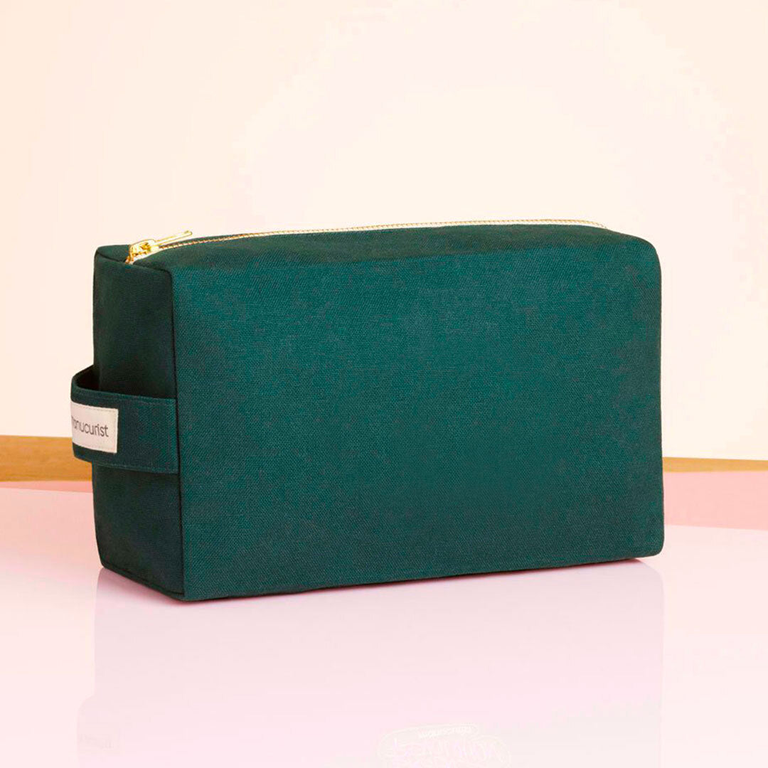 Le Large Green Bag de Manucurist-us est une pochette rectangulaire en coton biologique vert foncé, dotée d’une poignée latérale et d’une fermeture éclair dorée. Il est présenté sur une surface rose brillante avec un fond clair.