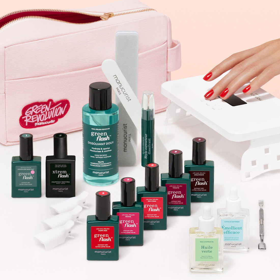 The Manucurist Super Ultimate Kit, featuring Green Flash gel polish, remover, cuticle oil, nail file, buffer, and a UV lamp, is displayed before a pink pouch as a hand with red nails uses the lamp for an ultra-glossy manicure.