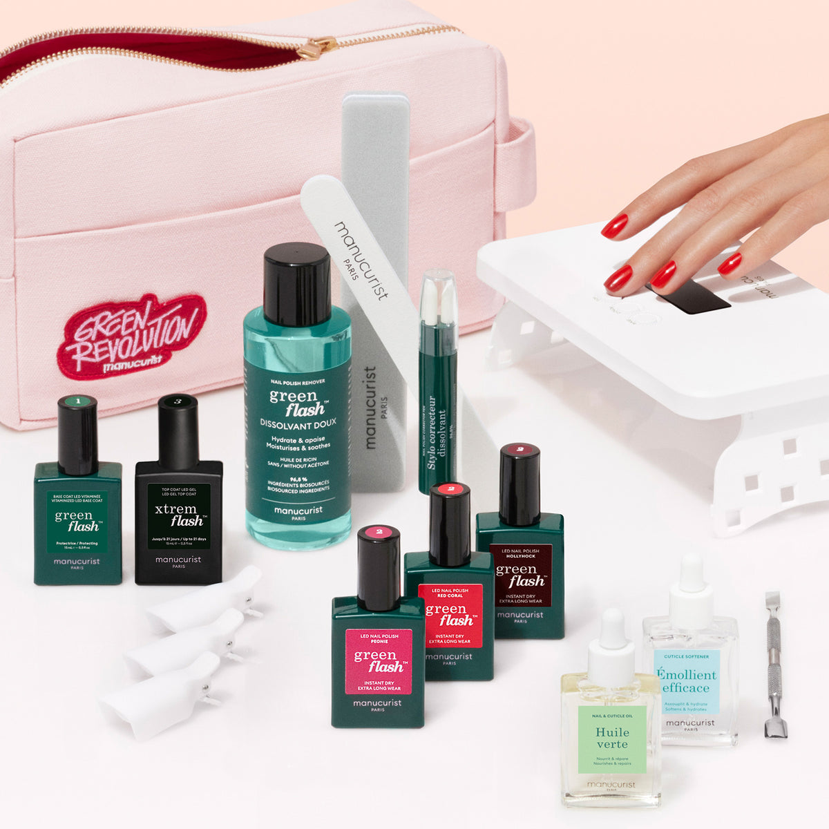 The Manucurist Ultimate Kit, featuring gel polish, remover, nail files, and Vitaminized Base Coat, is displayed before a pink pouch as a hand with red nails cures under a white LED lamp for an ultra-glossy manicure.