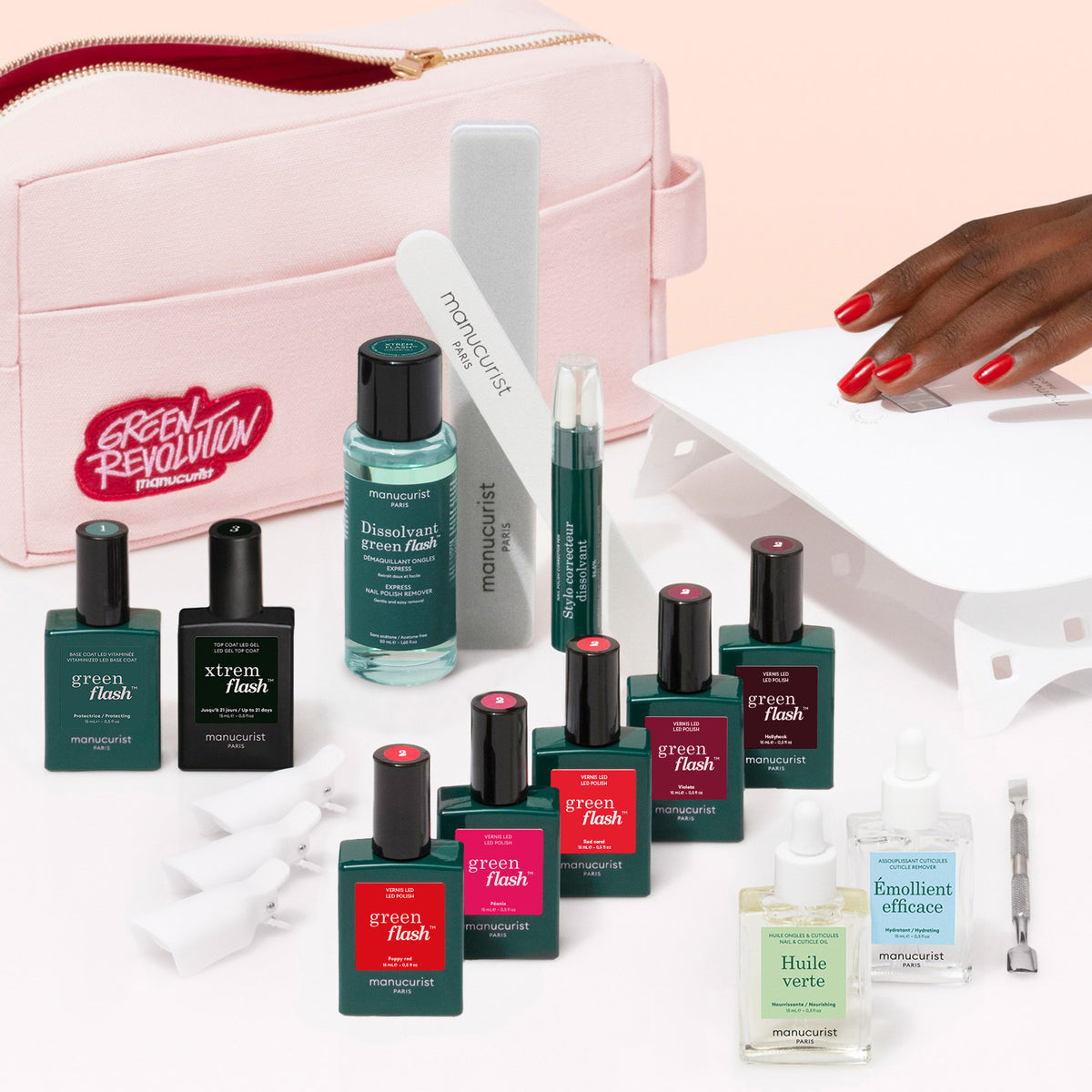 Le coffret Manucurist Super Ultimate Kit, comprenant le vernis Green Flash, du dissolvant, des huiles pour ongles, des limes et une base enrichie en vitamines avec une trousse rose. Une main aux ongles rouges révèle un fini ultra-brillant sous une lampe LED blanche.