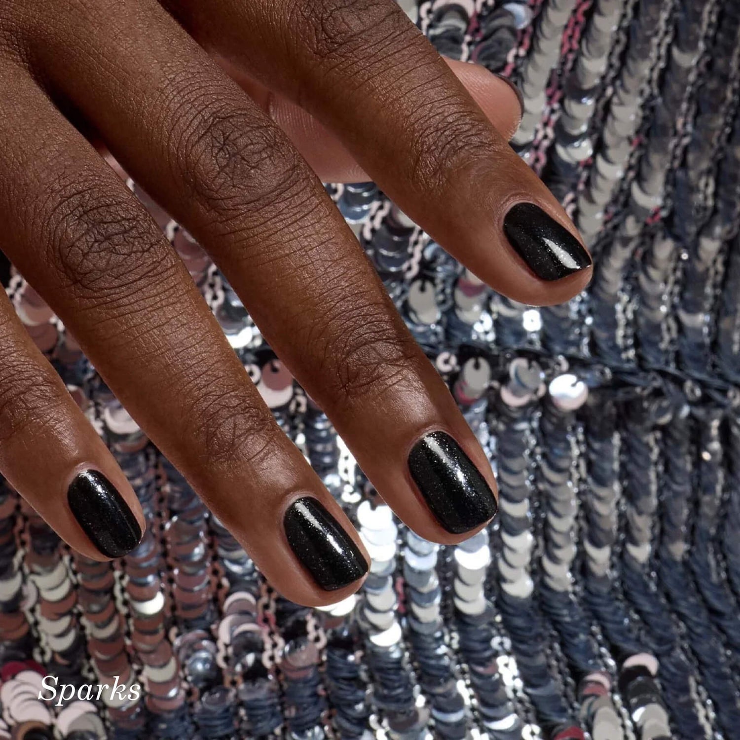A hand with short, neatly manicured nails painted glossy black with subtle sparkles rests on a sequined silver and black fabric.