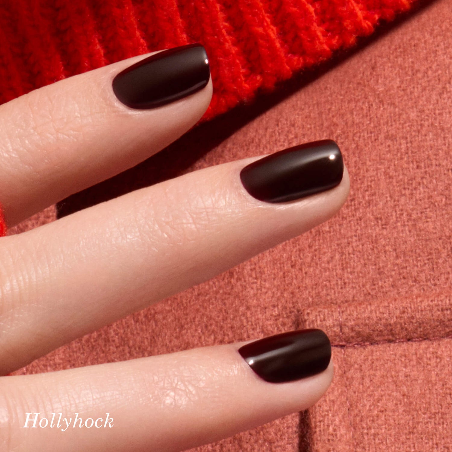 A close-up of a hand with short, glossy dark burgundy nails next to a green heart-shaped sticker labeled K4VN, with a red knit and quilted fabric in the background. The word Hollyhock appears in the lower left corner.