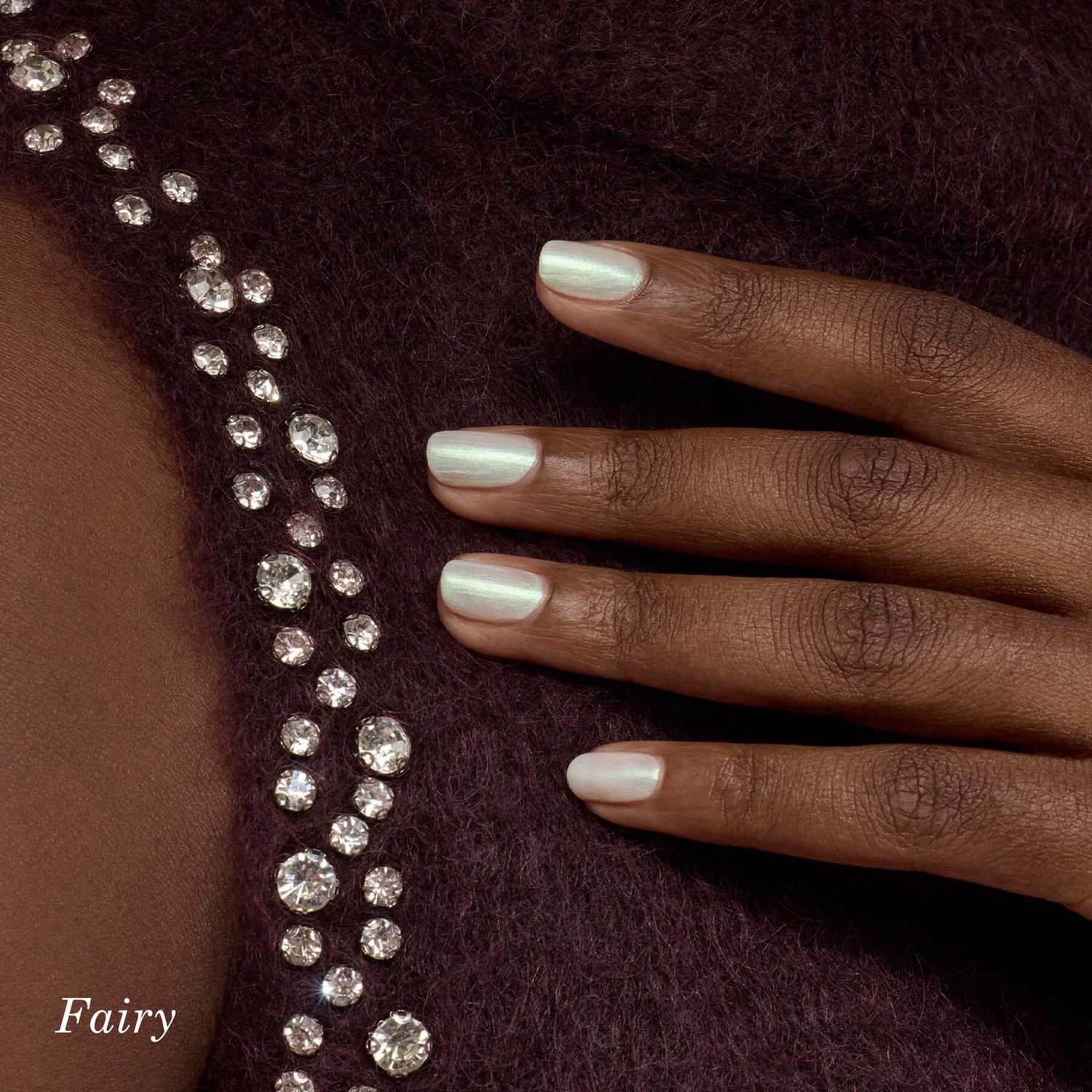 A hand with short, iridescent light pink nails rests on a dark purple, fuzzy fabric adorned with sparkling rhinestones. The word Fairy appears in the bottom left corner.