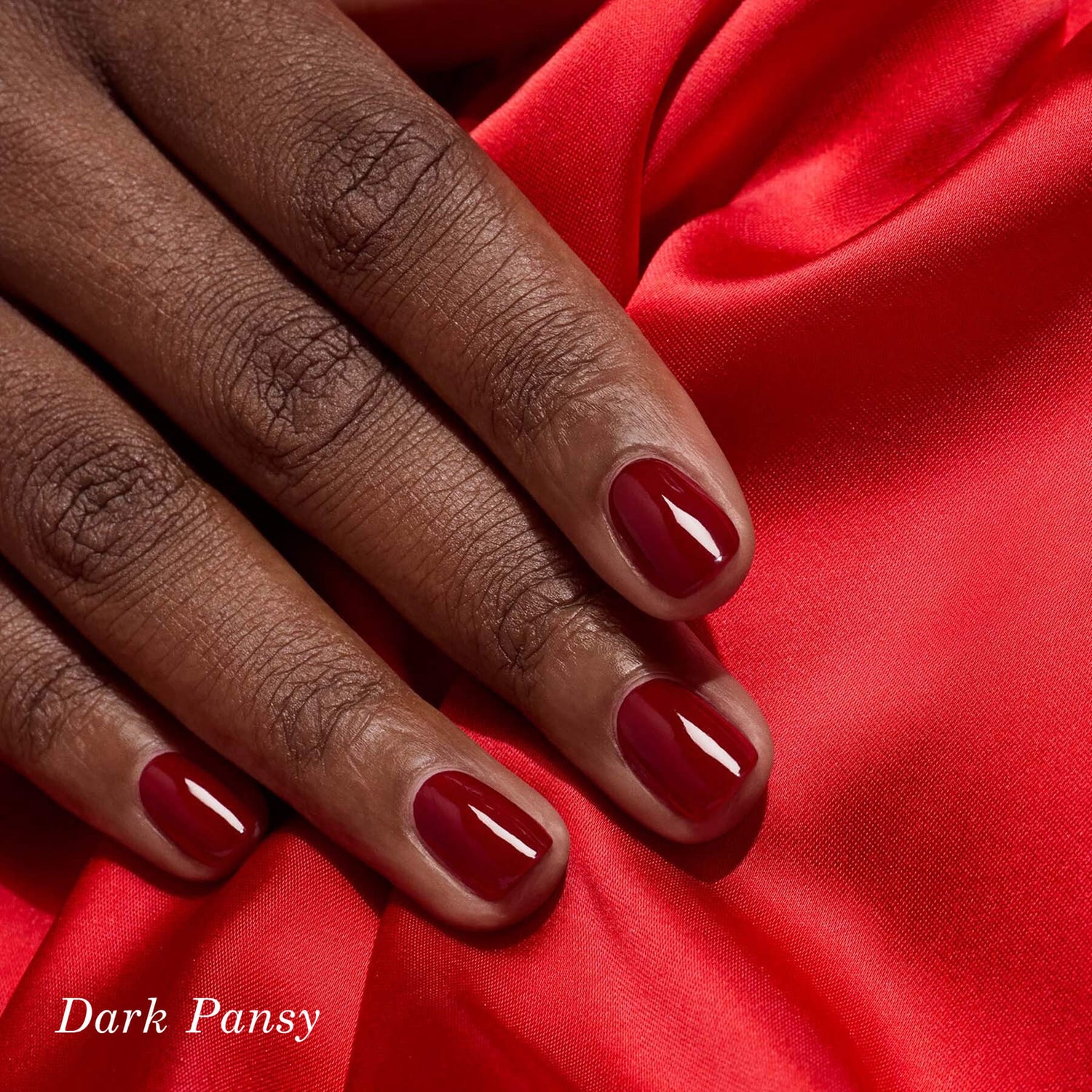 A close-up of a hand with dark skin resting on bright red fabric, featuring neatly manicured nails painted a glossy deep red shade. The text “Dark Pansy” appears in the bottom left corner.