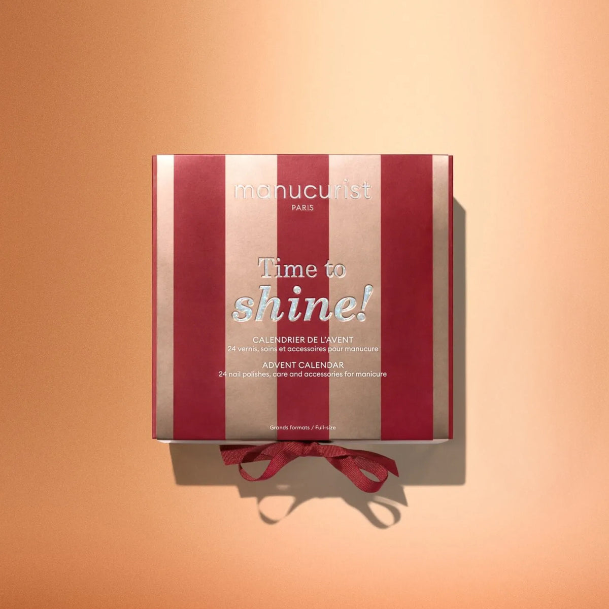 A red and gold Manucurist Advent Calendar Time to Shine! box with a ribbon, displayed on a peach background, featuring nail polishes, manicure accessories, and luxurious nail and hand care products like the innovative Active™ Shine Icy.