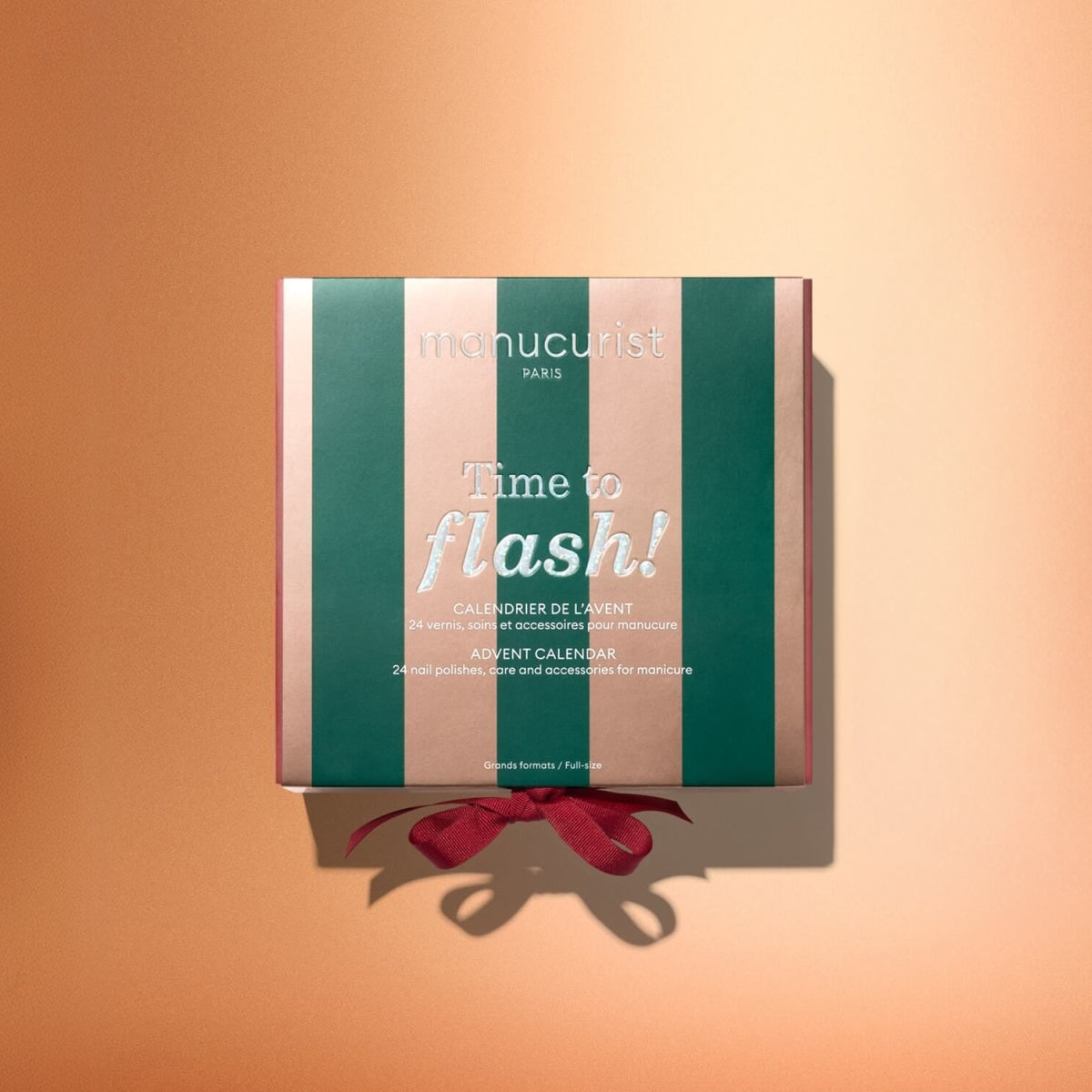 Centered against a peach background, the Manucurist Advent Calendar Time to Flash! features a square box adorned with green and pink stripes, the words “manucurist Paris Time to flash!” on the front, and is finished with a red ribbon—an ideal gift for anyone eager to explore manicure surprises or dive into the Green Flash collection.