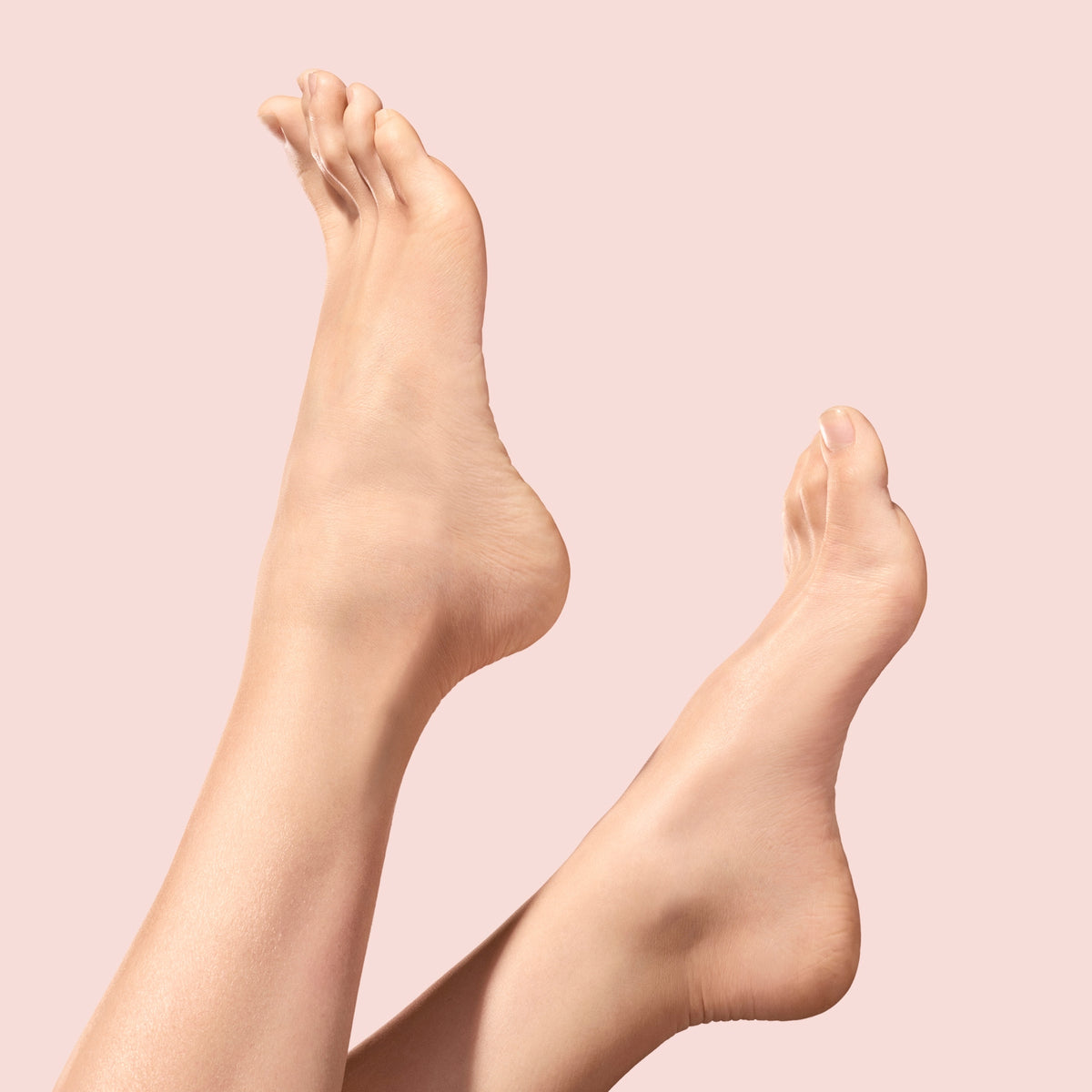 Two bare feet with smooth skin are pointed upward against a soft pink background, with toes slightly spread and ankles visible. The pose highlights the soles and curves of the feet.