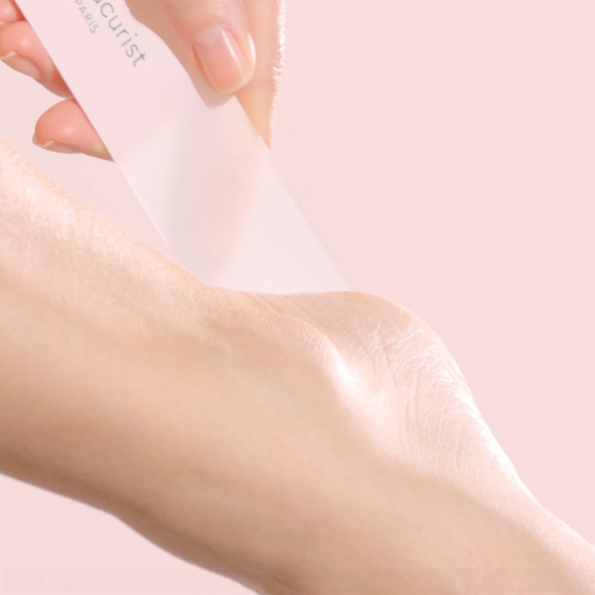 A hand gently files the heel of a foot with a translucent glass foot file against a soft pink background.