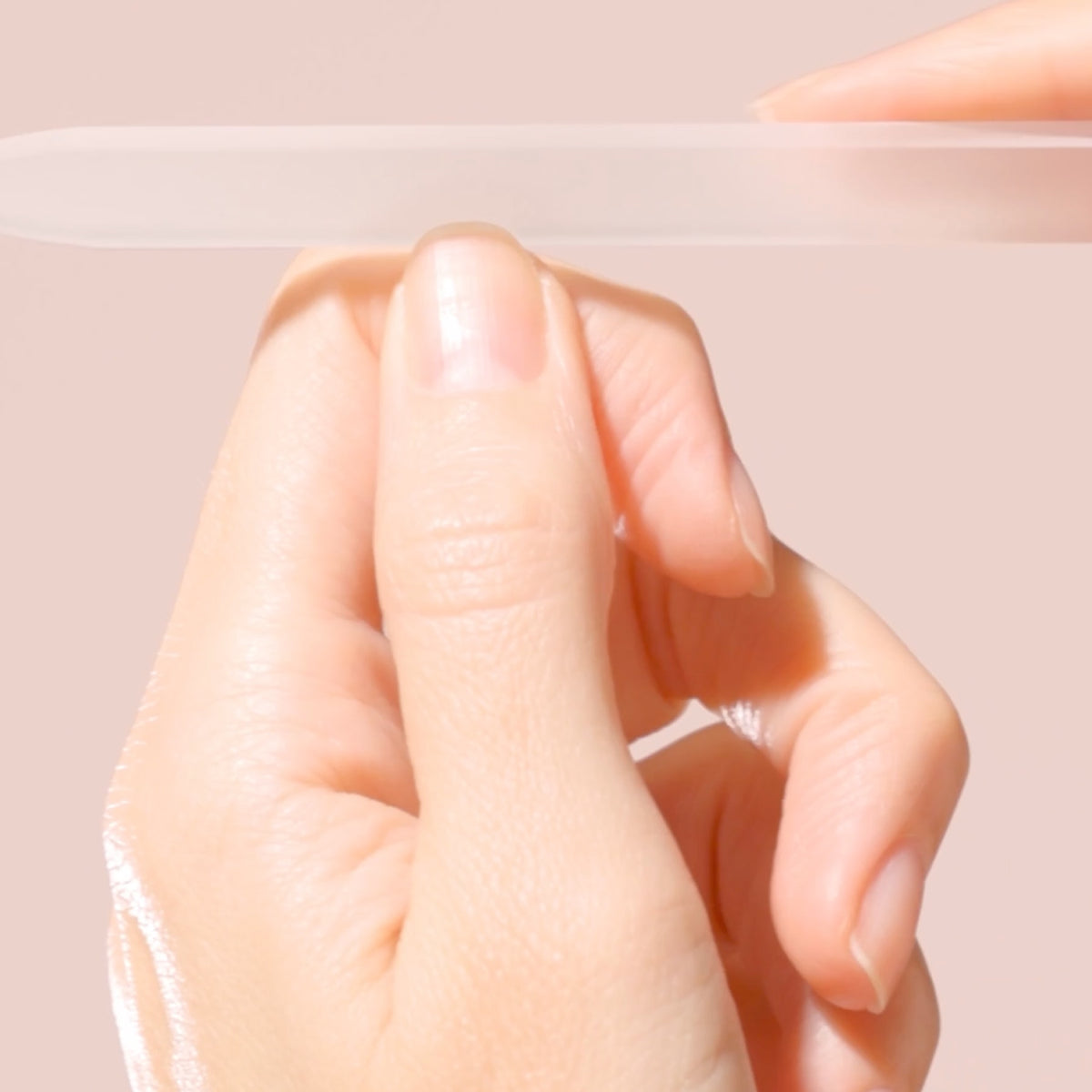 A close-up of a hand filing fingernails with a translucent nail file against a soft pink background. The hand has neatly manicured nails.