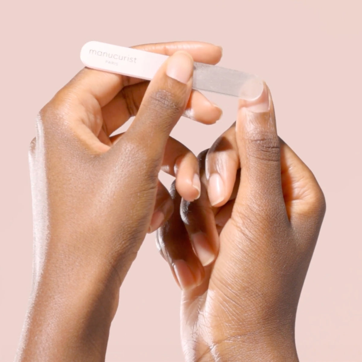 A person uses a nail file labeled manucurist to shape the nails on one hand, set against a soft pink background.