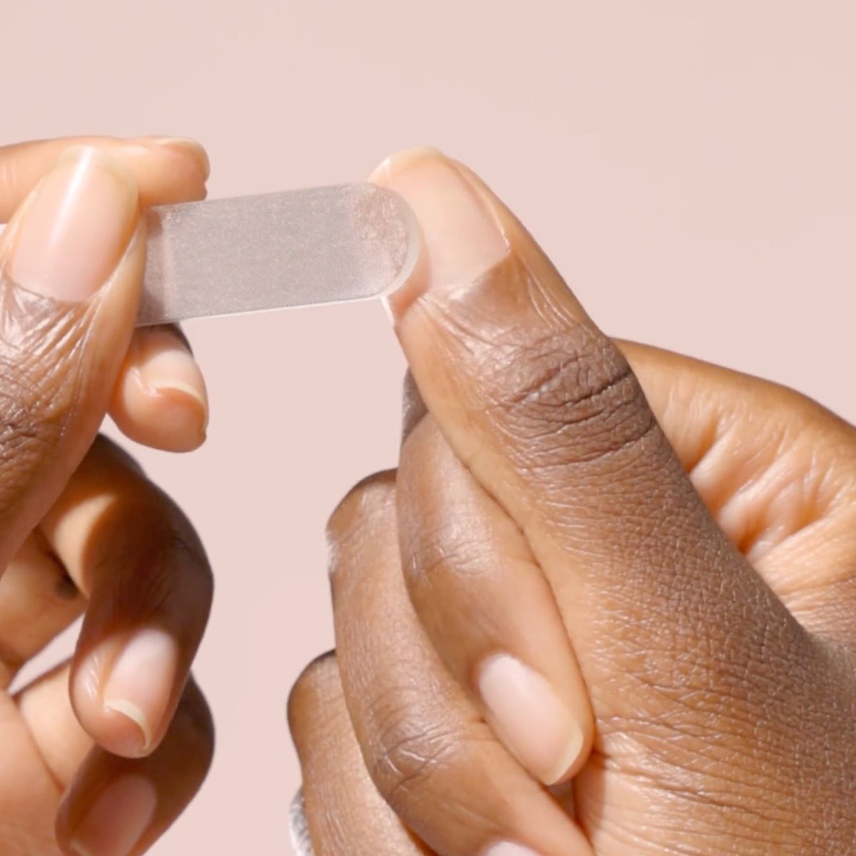 Close-up of two hands holding a small, clear adhesive bandage against a soft pink background.