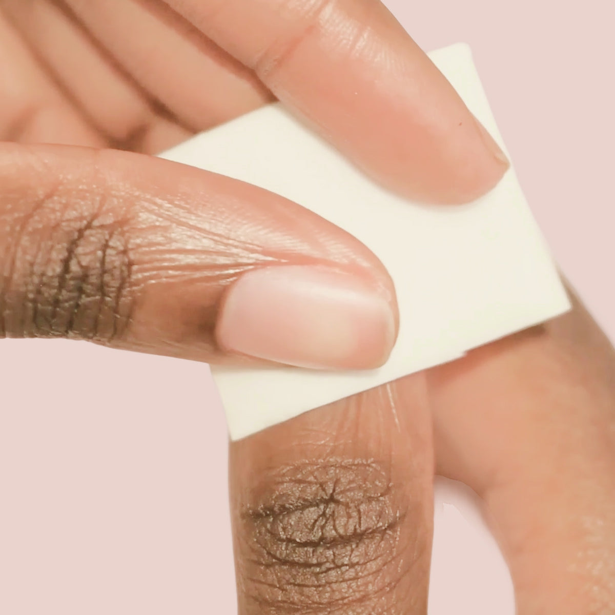 A close-up of a person using a white cotton pad to clean or remove nail polish from their fingernail against a pale pink background.