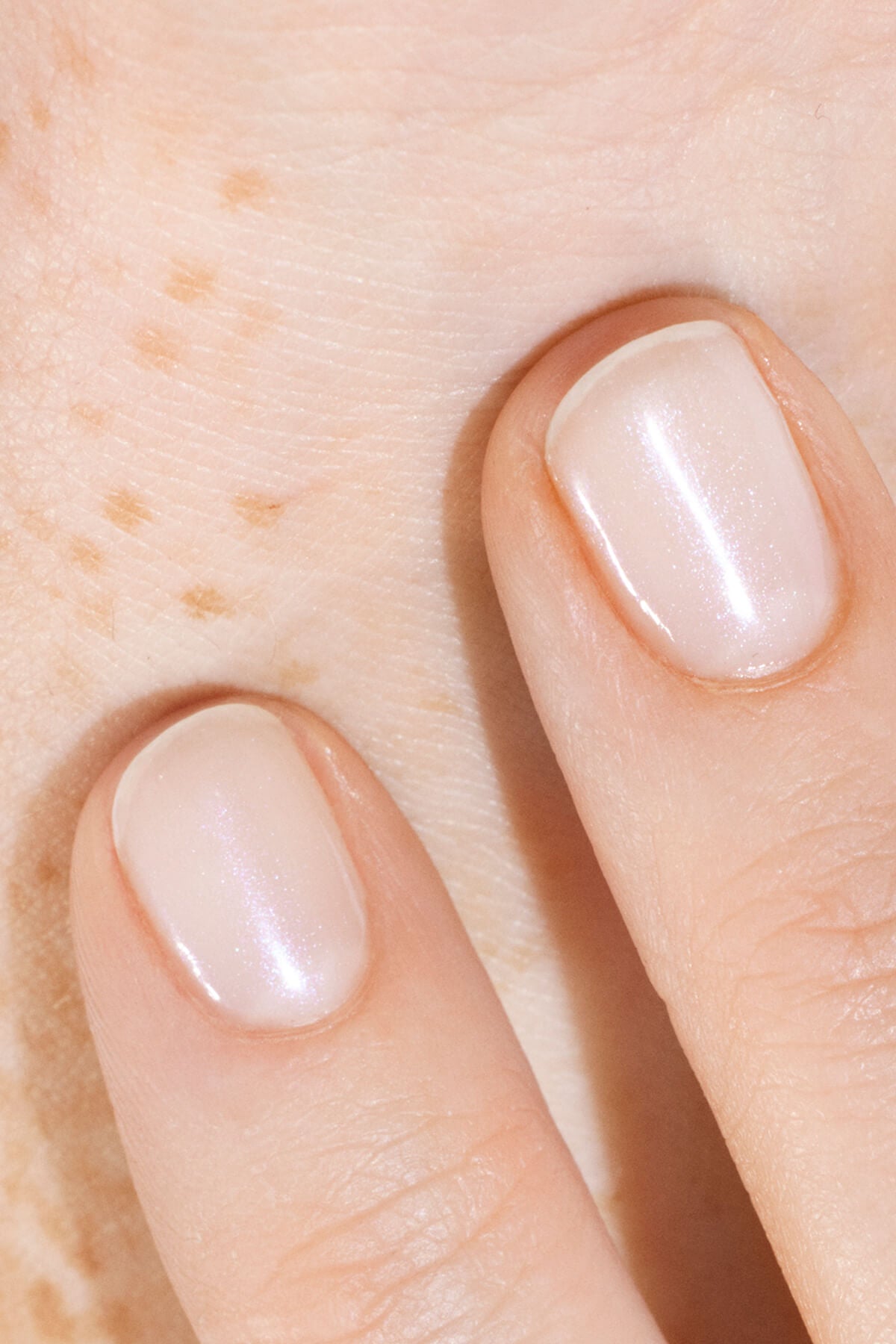 Gros plan sur deux doigts aux ongles courts et soignés, peints d’un vernis clair et nacré, reposant sur une peau claire où de légères taches de rousseur sont visibles.