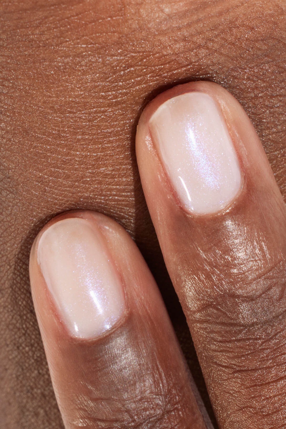 Gros plan sur deux doigts aux ongles soigneusement manucurés, vernis d’un rose clair transparent et scintillant, reposant sur une peau brune. Les ongles paraissent sains et brillants.