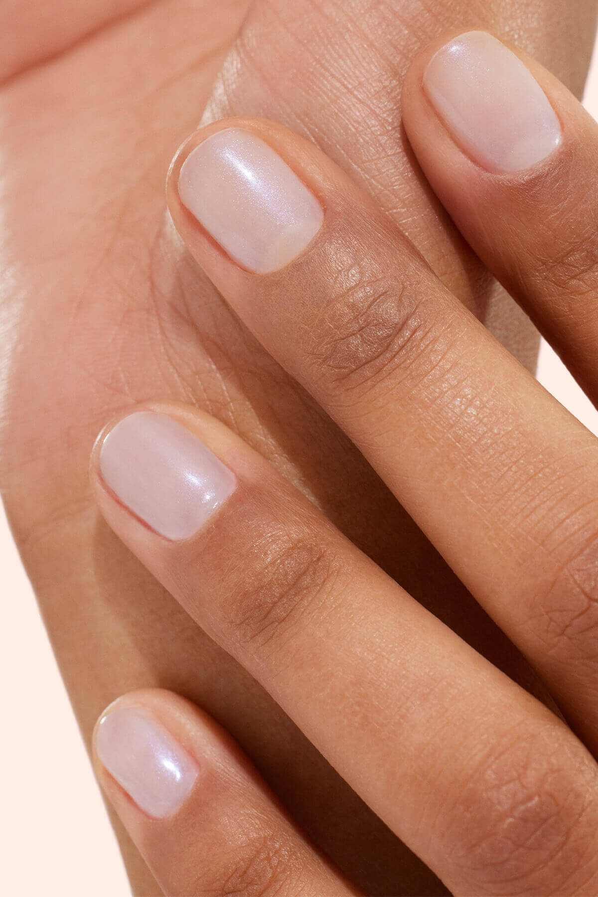 Gros plan sur une main aux ongles courts et soigneusement limés, vernis d'un rose pâle translucide, reposant délicatement sur la peau. Les ongles affichent un rendu net et naturel.
