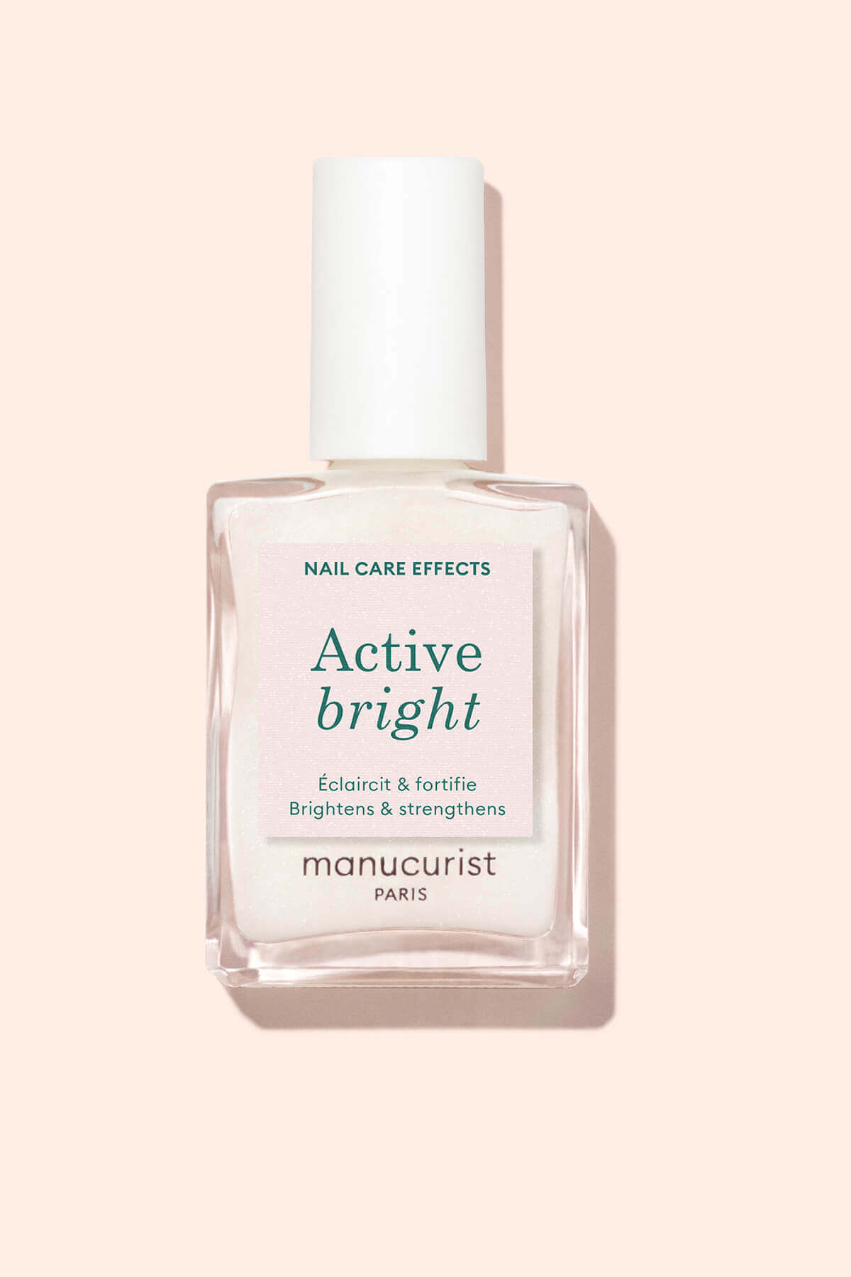 Un flacon en verre transparent de vernis Manucurist Active Bright avec un bouchon blanc, conçu pour illuminer et fortifier les ongles, posé sur un fond beige clair.