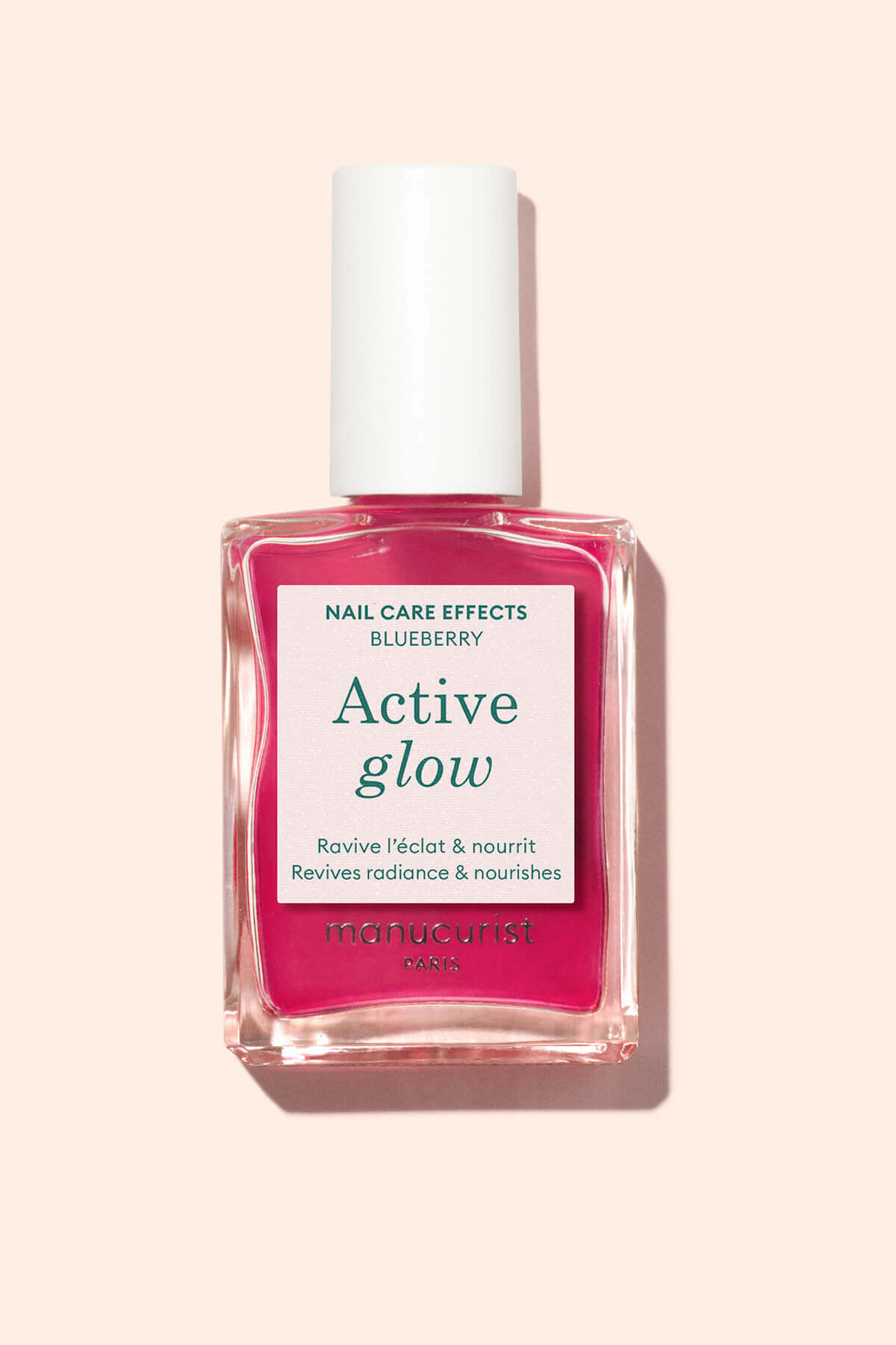 A bottle of Manucurist Paris Active Glow nail care polish with a white cap, pink liquid, and a label that reads Blueberry, Active glow, Revives radiance  nourishes on a light beige background.