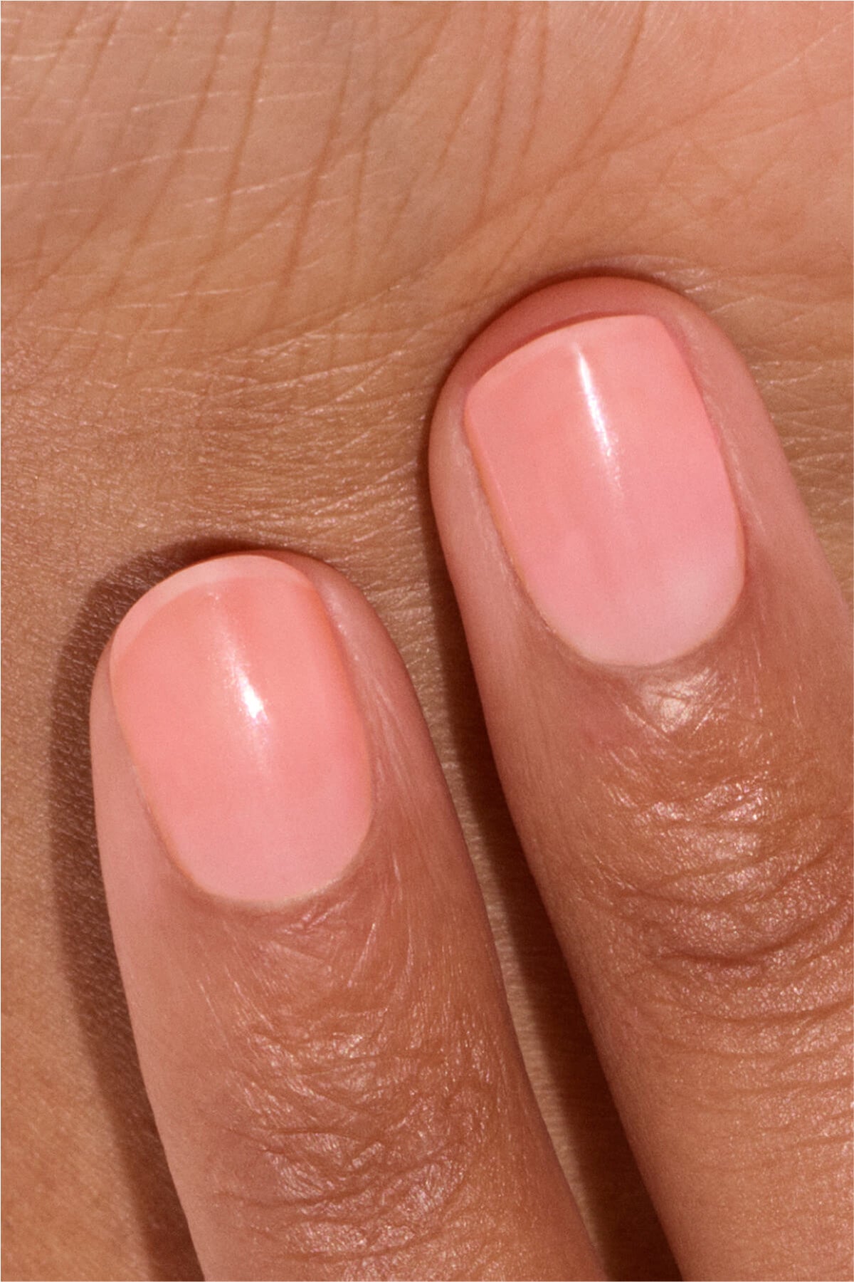 Deux doigts aux ongles soigneusement coupés, vernis d’un rose pâle brillant, sont présentés sur la peau d’une main. Les ongles paraissent sains et bien manucurés.