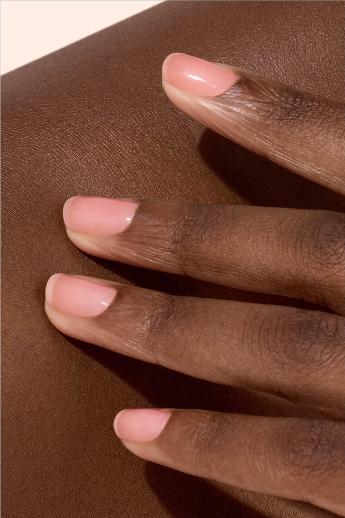 Gros plan d'une main aux ongles naturels et soignés, délicatement posée sur une peau lisse brun foncé. Les ongles sont courts et brillants, avec une douce nuance rose.