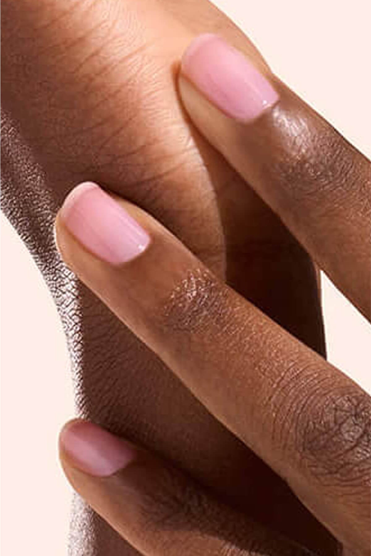 Gros plan d’une main aux ongles soigneusement manucurés, vernis d’un rose doux, reposant délicatement sur un avant-bras à la peau de teinte moyenne à foncée. Le fond est clair et uni.