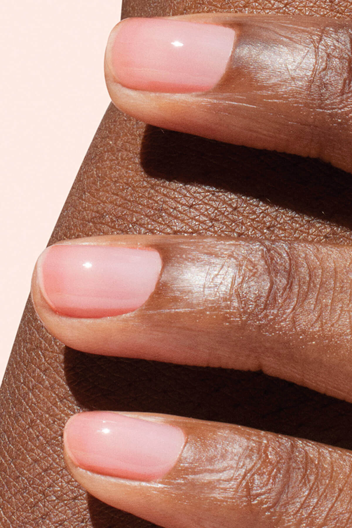 Gros plan sur trois doigts aux ongles soigneusement manucurés, vernis d’un rose pâle brillant, reposant sur une peau brun foncé.