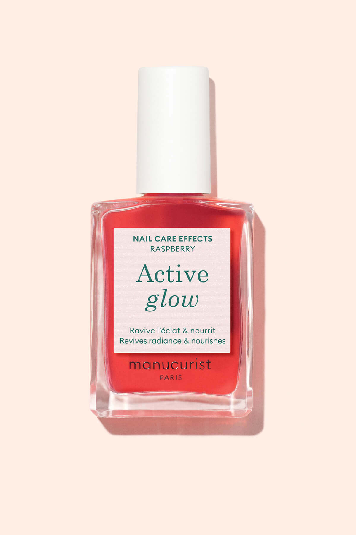 A clear glass bottle of Manucurist Paris Active Glow nail care polish with a white cap, filled with a bright coral-pink liquid, photographed on a light peach background.