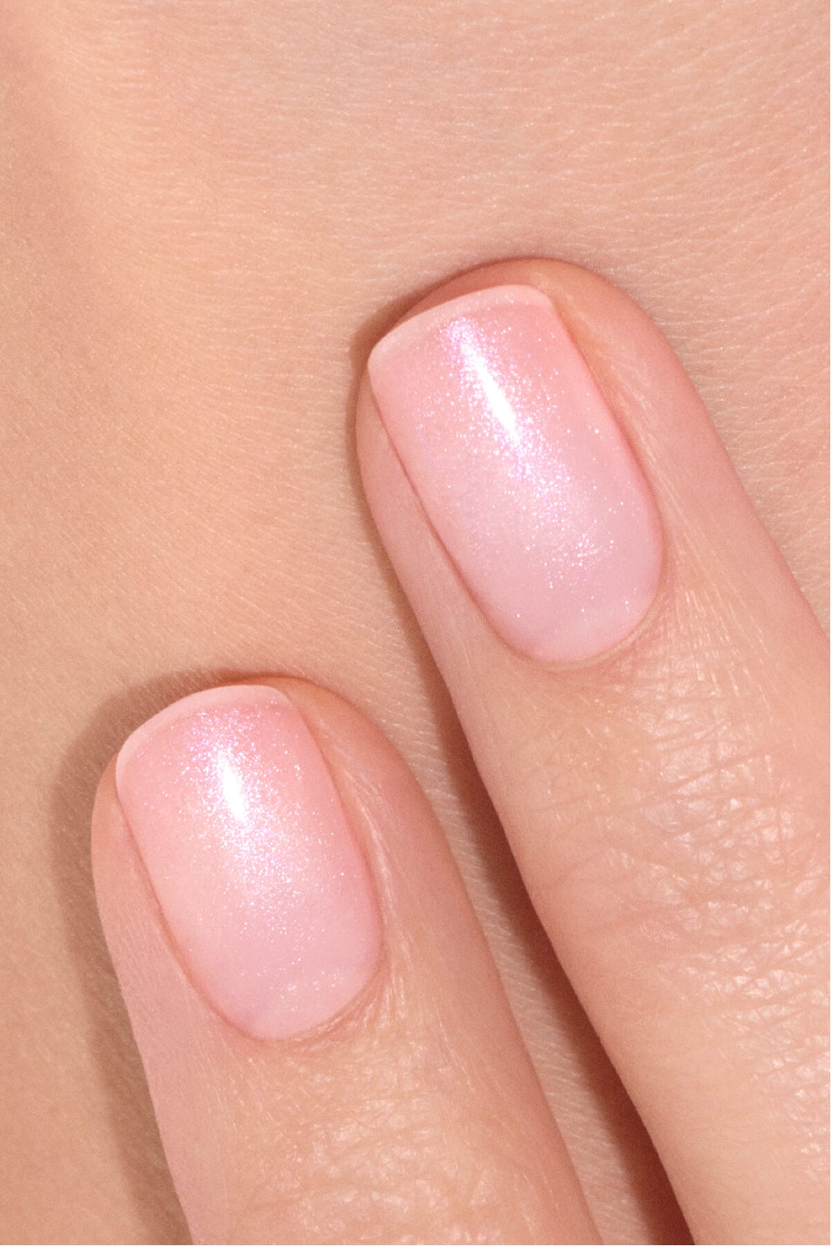 Gros plan sur deux doigts aux ongles soigneusement manucurés, peints d’un vernis rose pâle scintillant, posés contre la peau.