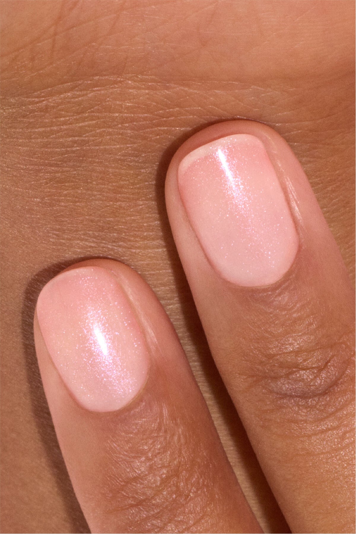 Gros plan sur deux doigts aux ongles courts, soigneusement manucurés et peints d’un vernis à ongles rose pâle scintillant, posés sur un fond de peau brune.