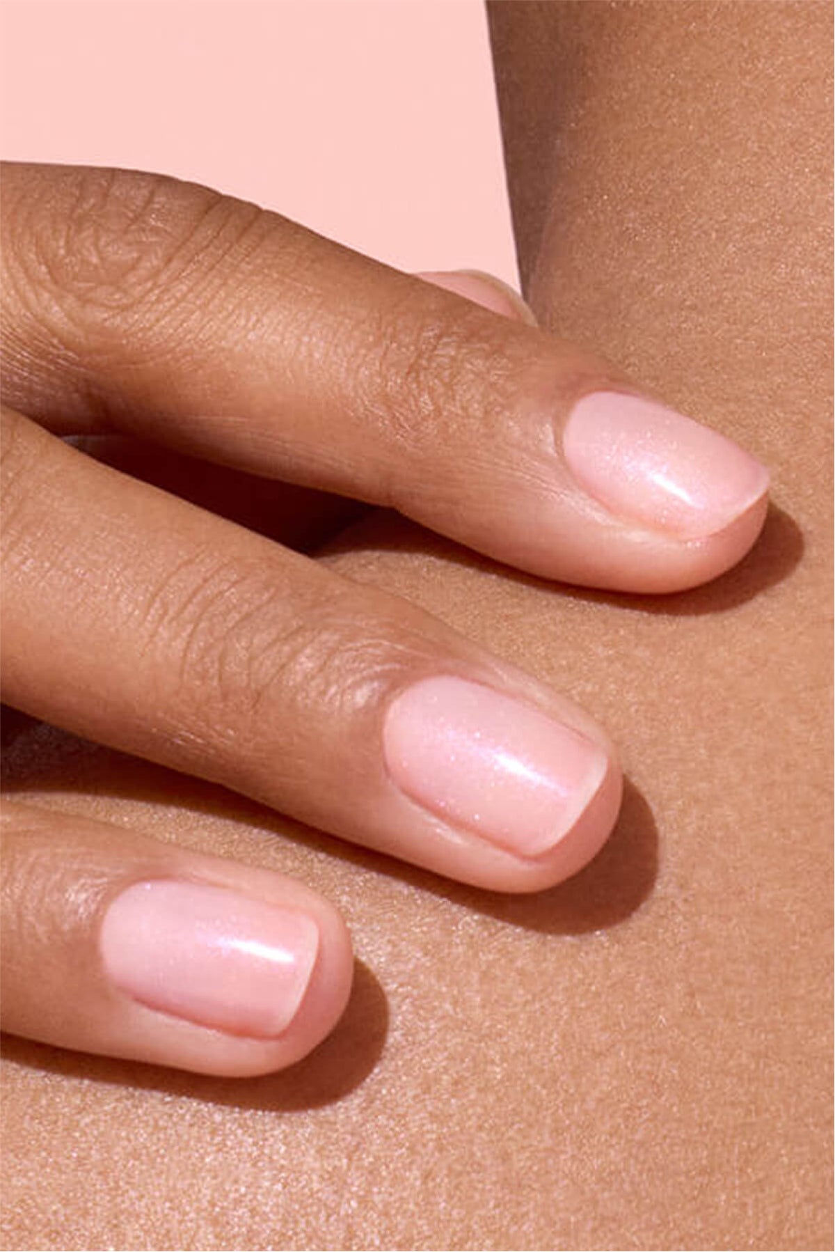 Gros plan d'une main aux ongles soigneusement manucurés d'un rose clair et doux, délicatement posée sur la peau d'une personne sur fond rose.