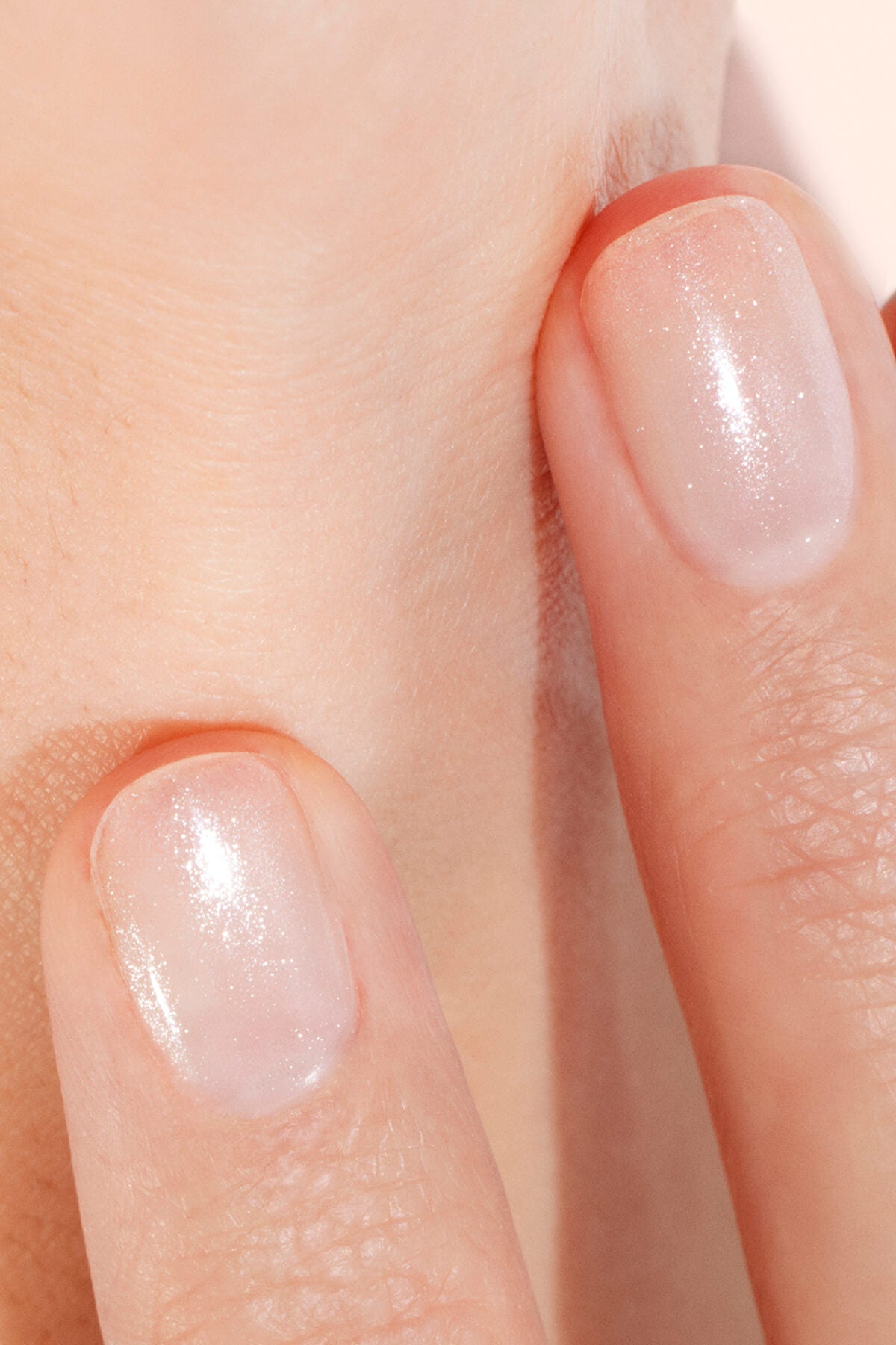 Gros plan sur deux doigts aux ongles courts et soignés, vernis d'un rose pâle transparent et scintillant, touchant délicatement une peau claire.