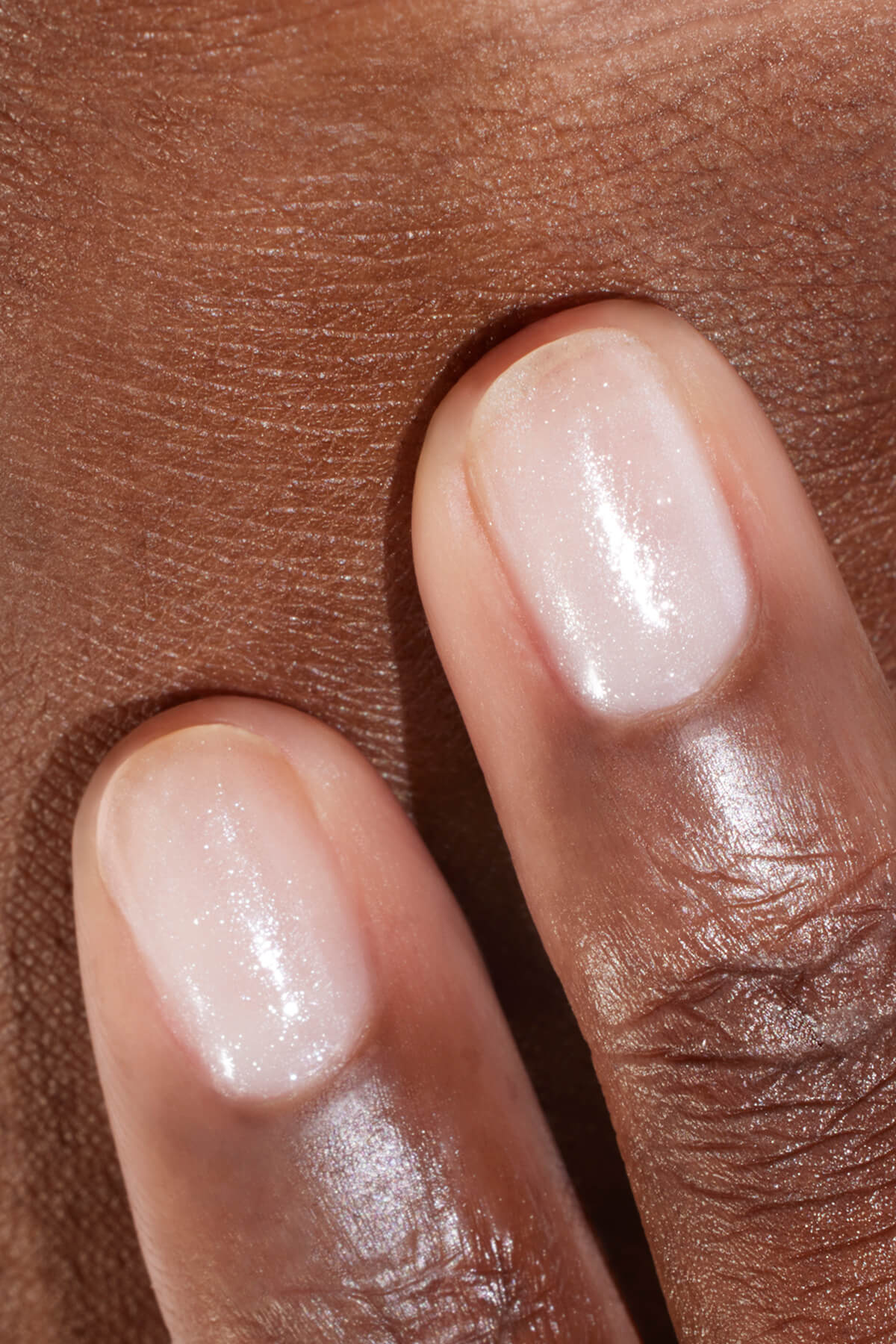 Gros plan sur deux doigts aux ongles courts et impeccablement manucurés, vernis d'une teinte rose clair transparente et scintillante, posés sur une peau mate.