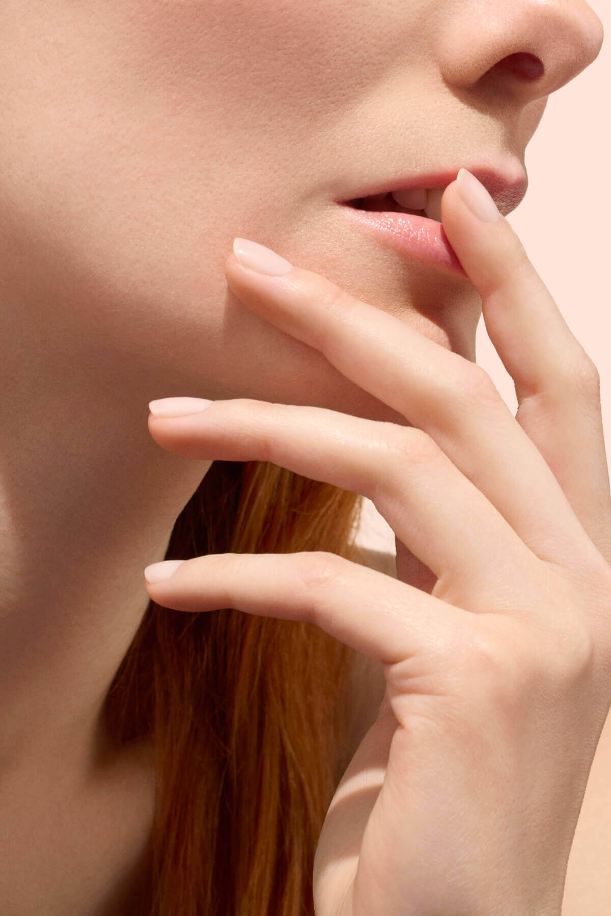 Gros plan sur le visage et la main d'une femme, ses doigts effleurant délicatement ses lèvres. Elle a la peau claire, un teint lisse et un vernis à ongles nude et naturel. Le fond est doux et neutre.