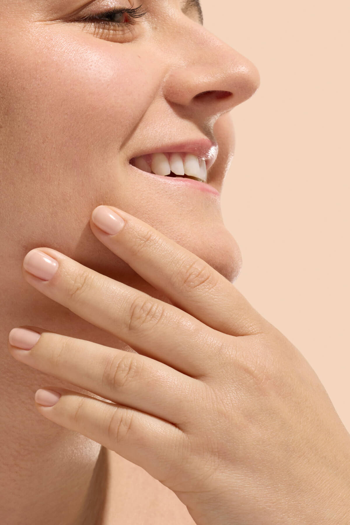 Gros plan sur une femme souriante, montrant le bas de son visage et sa main touchant son menton. Sa peau paraît lisse et ses ongles sont courts et naturels. L'arrière-plan est d'une couleur beige doux.