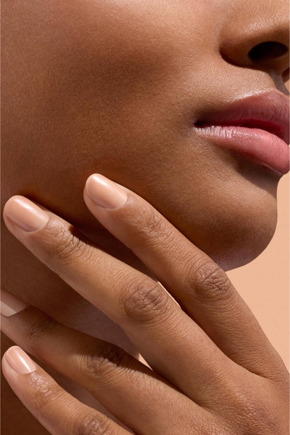 Gros plan sur le bas du visage et le menton d’une personne, dont les doigts touchent délicatement la mâchoire. La personne a la peau lisse, un maquillage naturel et des ongles soigneusement manucurés.