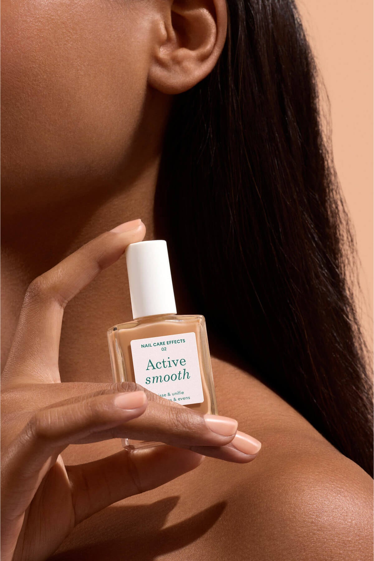 Une personne aux longs cheveux bruns tient près de son visage un petit flacon de soin pour les ongles étiqueté Active Smooth, mettant en valeur le produit contre son épaule nue sur fond beige.